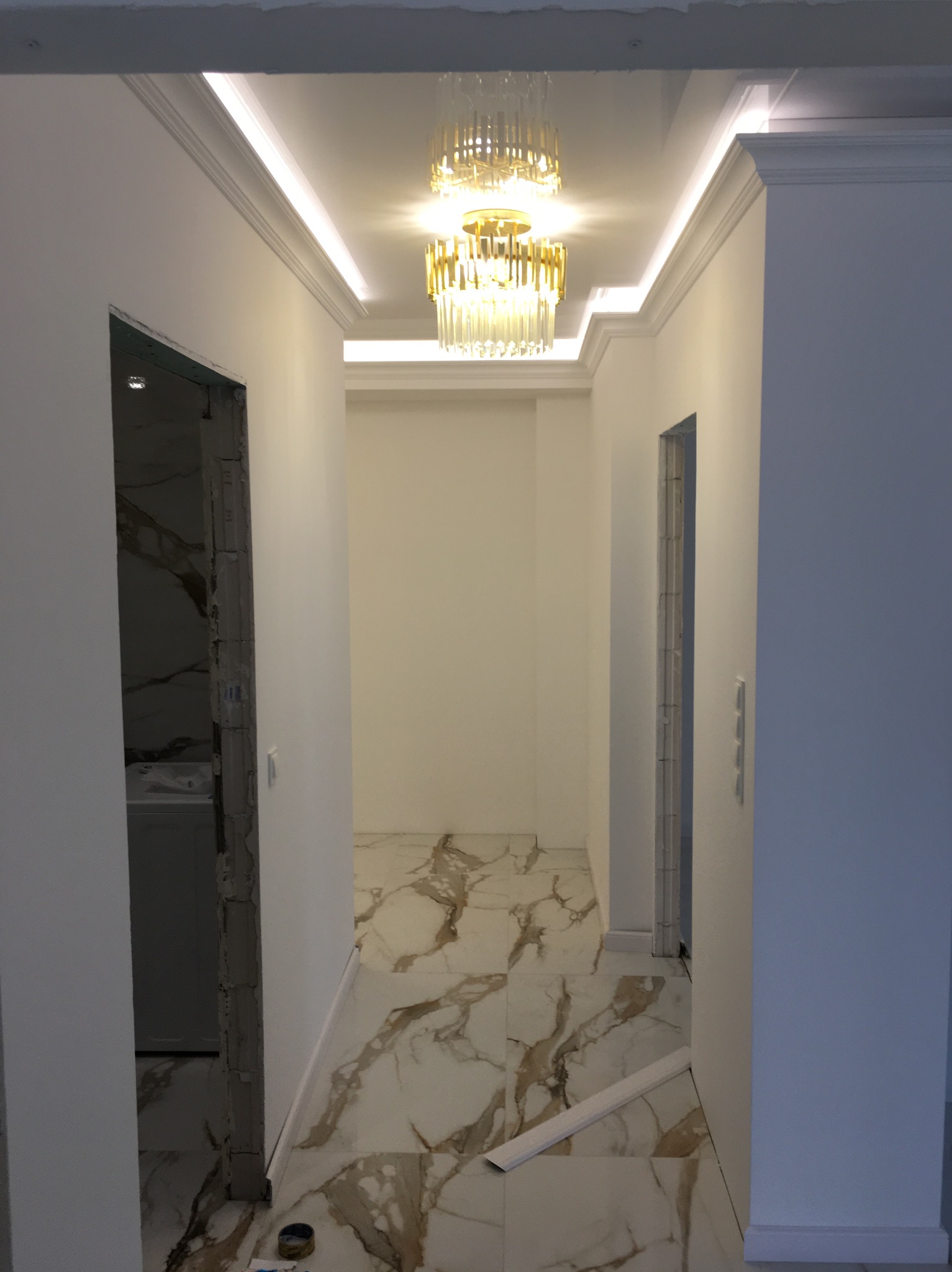 Korytarz w trakcie prac wykończeniowych: położona marmurowa podłoga, zamontowane listwy przypodłogowe, sufit z oświetleniem LED i ozdobnym żyrandolem, widoczne niedokończone futryny drzwiowe.