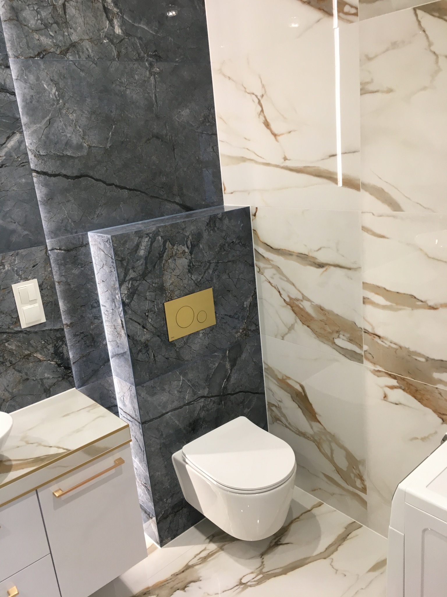 Nowoczesna łazienka z wiszącą toaletą, marmurowymi płytkami w odcieniach szarości i bieli z brązowymi żyłami, oraz złotymi detalami na przycisku spłuczki i szafce umywalkowej.