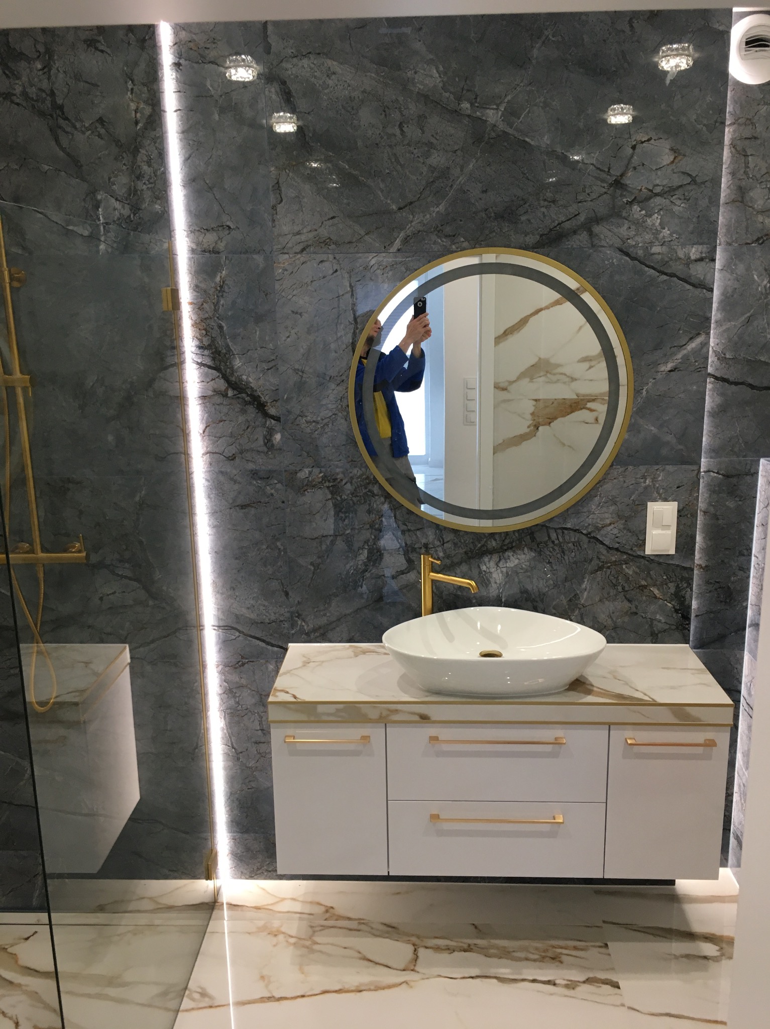 Nowoczesna łazienka z marmurowymi płytkami, okrągłym lustrem w złotej ramie, białą szafką pod umywalkę z złotymi uchwytami i oświetleniem LED w pionowym pasie.