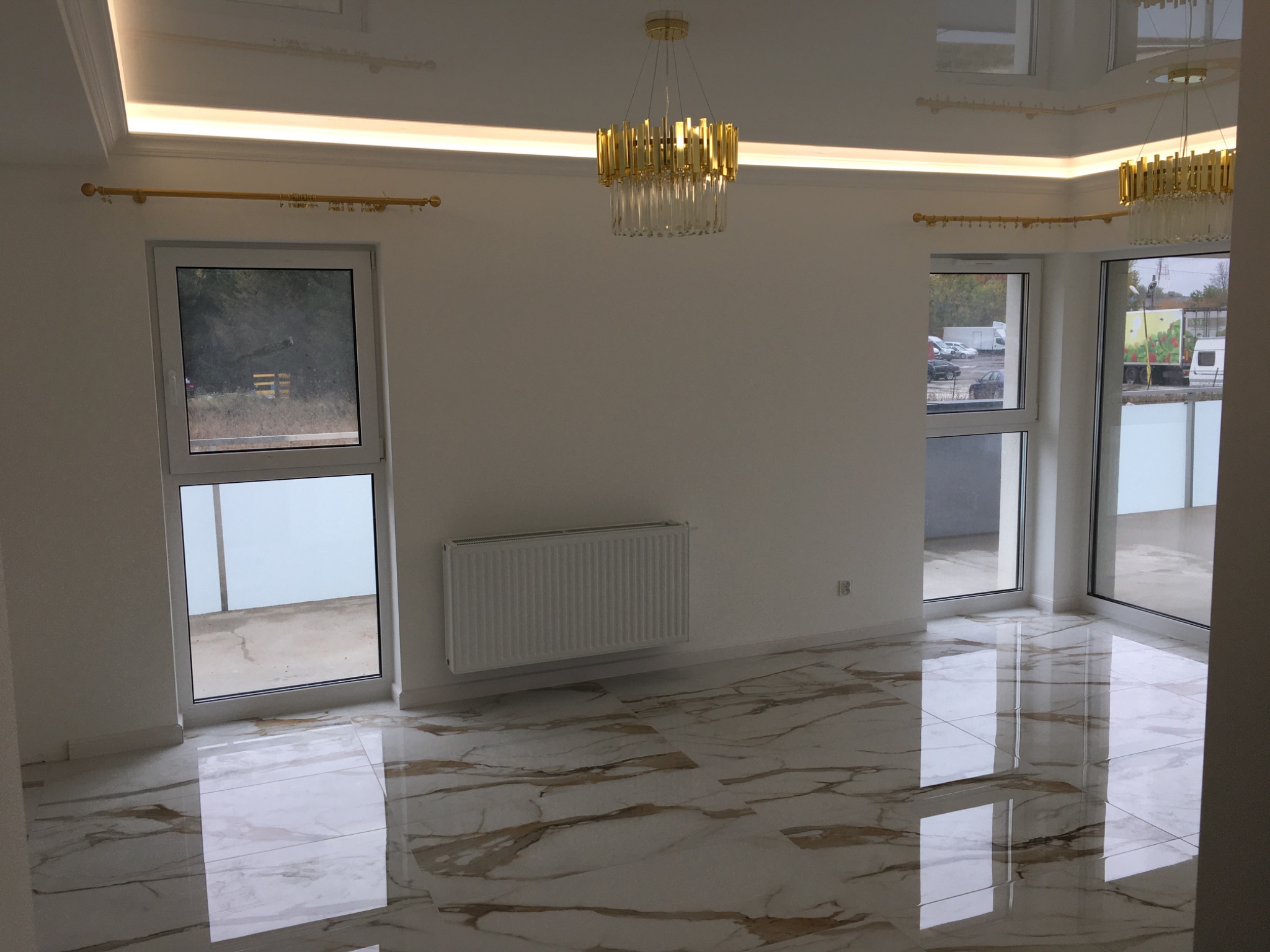 Puste, jasne wnętrze z marmurową podłogą, złotym żyrandolem i karniszami, grzejnikiem oraz dużymi oknami z widokiem na zewnątrz.