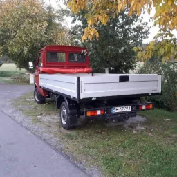 Czerwony samochód dostawczy z otwartą, pustą skrzynią ładunkową, zaparkowany na poboczu drogi wśród drzew z żółtymi liśćmi, widok z tyłu, tablica rejestracyjna SH 6779A.