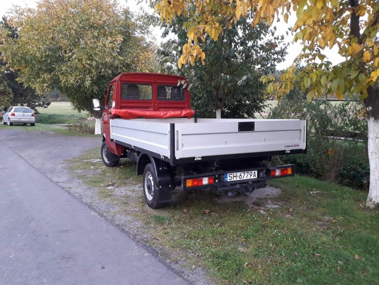 Czerwony samochód dostawczy z otwartą, pustą skrzynią ładunkową, zaparkowany na poboczu drogi wśród drzew z żółtymi liśćmi, widok z tyłu, tablica rejestracyjna SH 6779A.