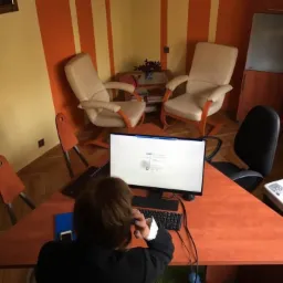 Osoba siedząca przy trójkątnym biurku w biurze w Toruniu, z widocznym monitorem, klawiaturą, telefonem przy uchu, drukarką i dwoma fotelami w tle.