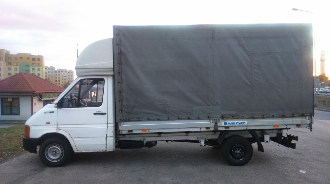 Biały bus z plandeką zaparkowany na ulicy, widoczny z boku na tle budynków mieszkalnych i kiosku.