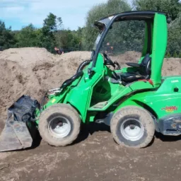 Zielona miniładowarka Avant 635 na placu budowy, z hałdą piasku w tle i pracownikami w oddali.