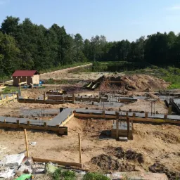 Wylane fundamenty pod dom jednorodzinny z drewnianym szalunkiem w otoczeniu drzew i małego stawu.