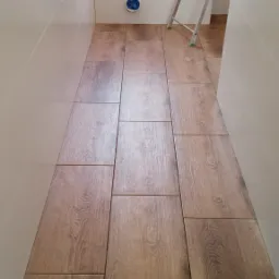 Ułożona podłoga z płytek imitujących drewno w wąskiej łazience z białą glazurą i drabinką aluminiową w tle.