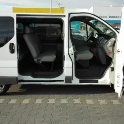 Biały van z otwartymi drzwiami przesuwnymi i przednimi, widoczne wnętrze z szarymi fotelami na tle parkingu przed sklepem.