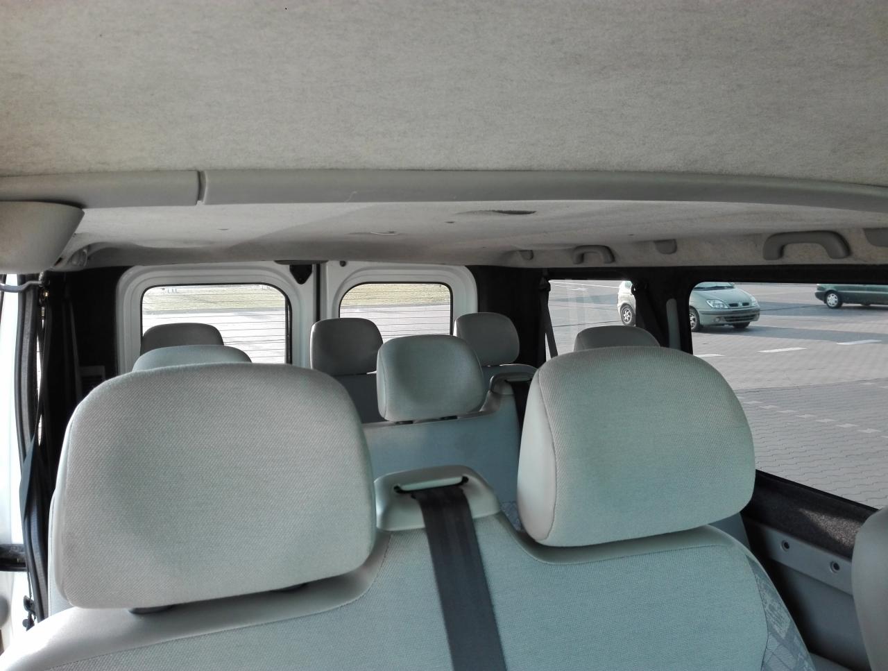 Wnętrze busa z jasnymi siedzeniami, widok z tyłu na zagłówki i pasy bezpieczeństwa, przez okna widoczne inne samochody na parkingu.