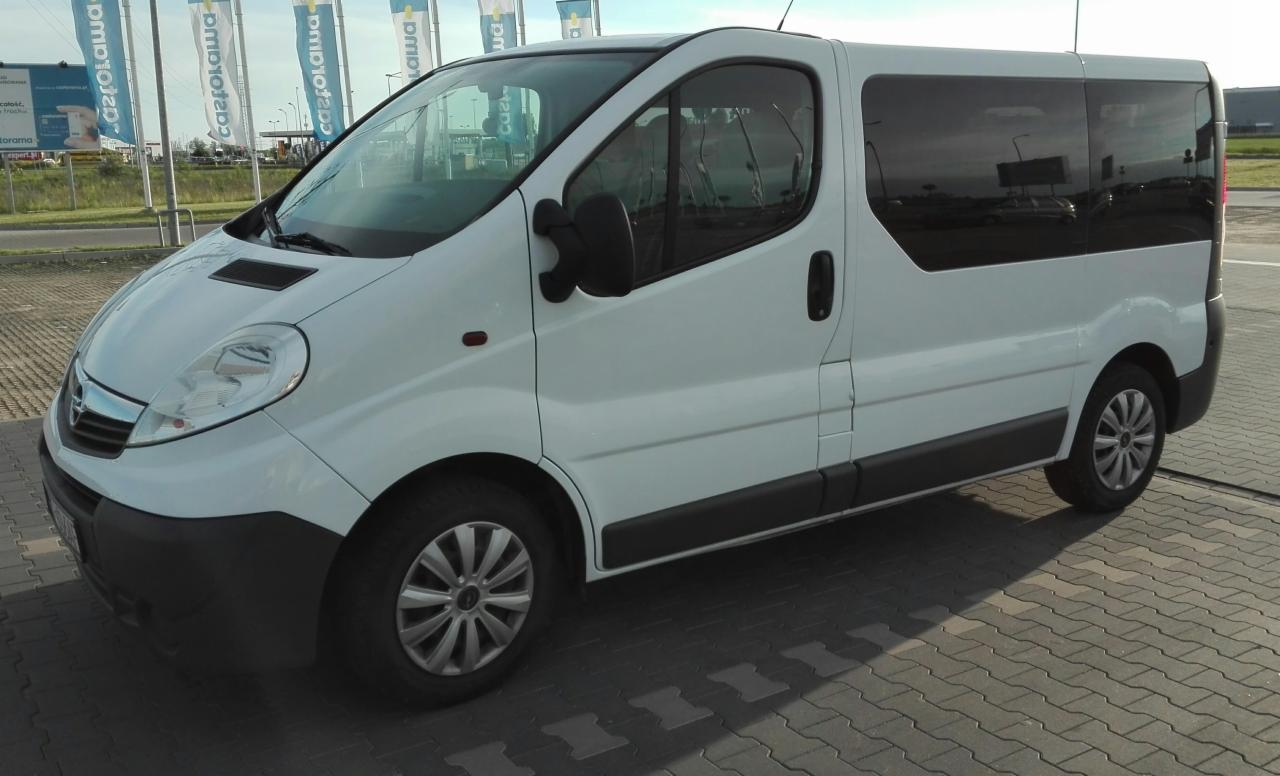 Biały Opel Vivaro z przyciemnianymi szybami, zaparkowany na kostce brukowej w pobliżu drogi i słupów reklamowych.