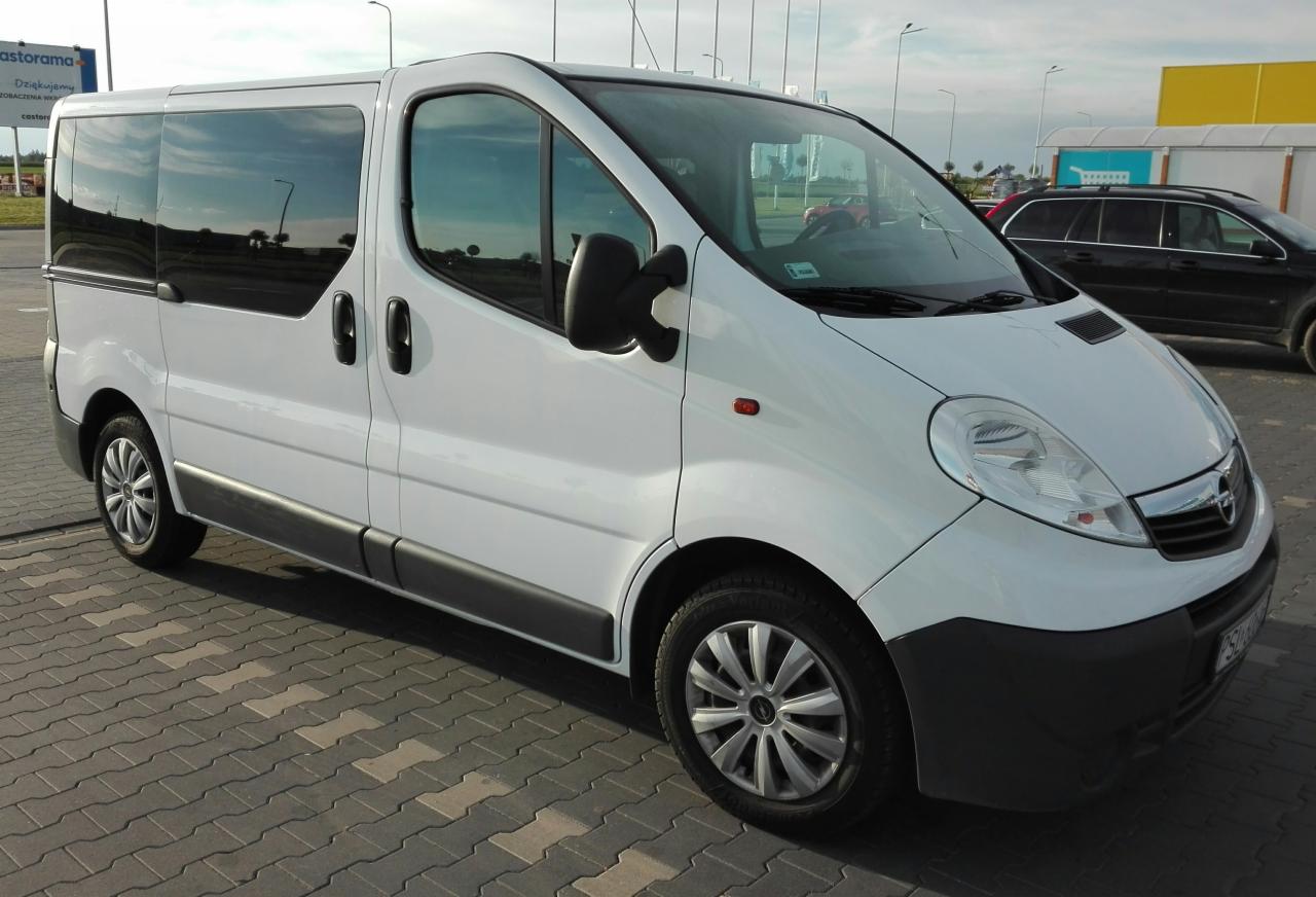 Biały van Opel Vivaro z przesuwnymi drzwiami, zaparkowany na brukowanej powierzchni, z widocznym odbiciem nieba w szybach.