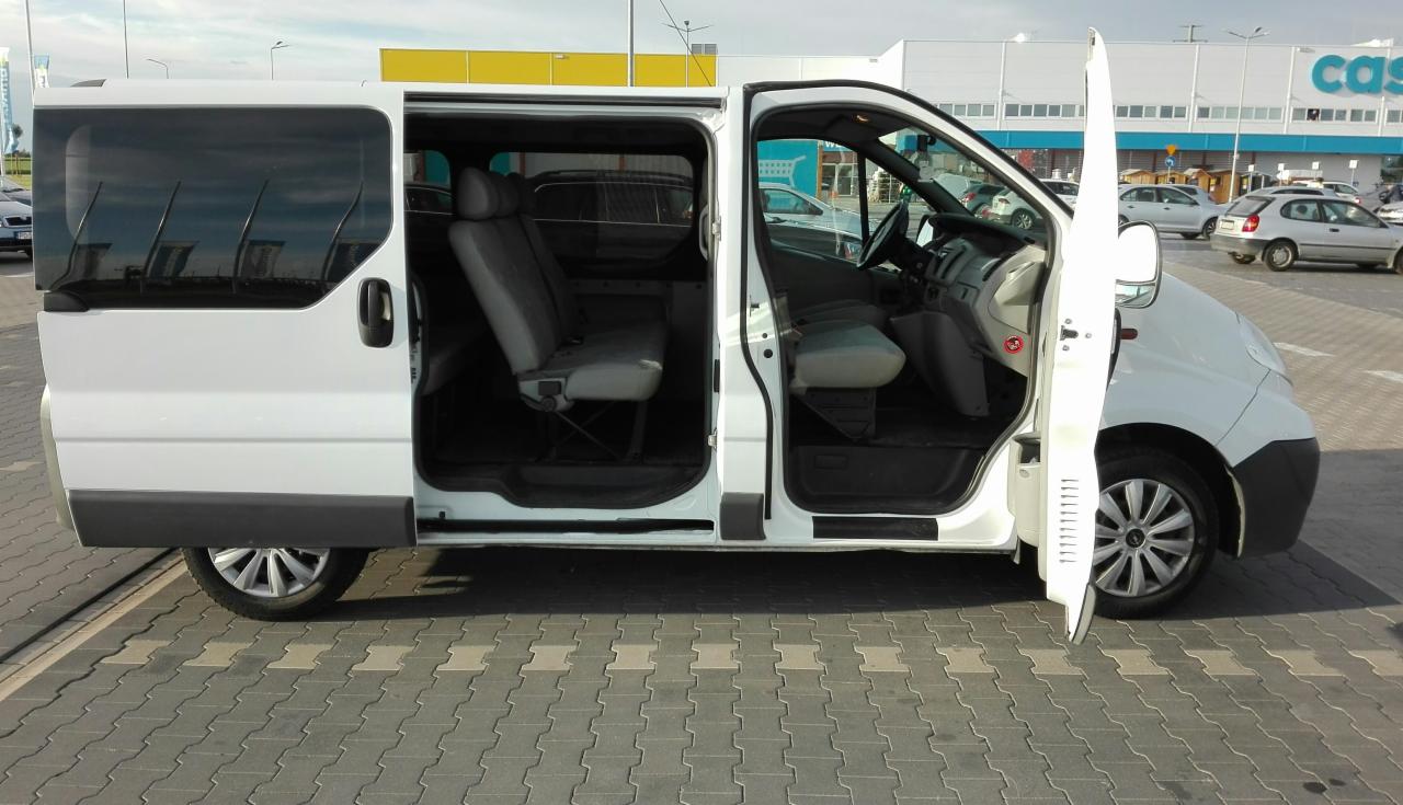 Biały van z otwartymi drzwiami przesuwnymi i przednimi, widoczne wnętrze z szarymi fotelami na tle parkingu przed sklepem.