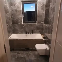 Wykończona łazienka z szarymi marmurkowymi płytkami, białą wanną z czarną armaturą, podwieszaną toaletą i oknem.