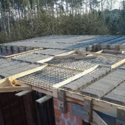 Wieniec stropowy w trakcie wykonywania, z widocznym deskowaniem i zbrojeniem, bloczki betonowe ułożone na stropie, w tle las.