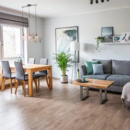 Jasne, przestronne wnętrze mieszkania z drewnianą podłogą, szarym narożnikiem z turkusowymi poduszkami, drewnianym stołem jadalnym i wiszącymi lampami z żarówkami Edisona.