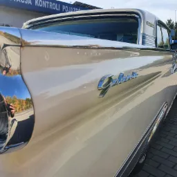 Fragment odrestaurowanego, kremowego Forda Galaxie z 1959 roku w Dąbrowie Chełmińskiej, z widocznym chromowanym reflektorem i napisem 'Galaxie', stojącego na kostce brukowej.