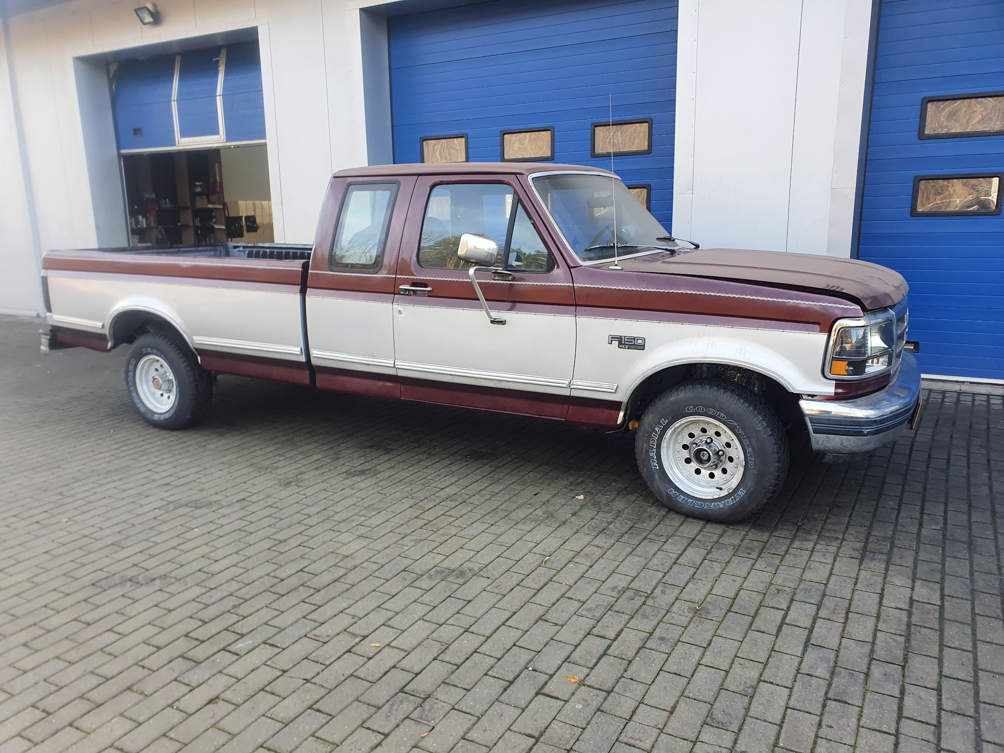 Dwukolorowy pickup Ford F-150 z lat 90. zaparkowany na kostce brukowej przed warsztatem z otwartą bramą.