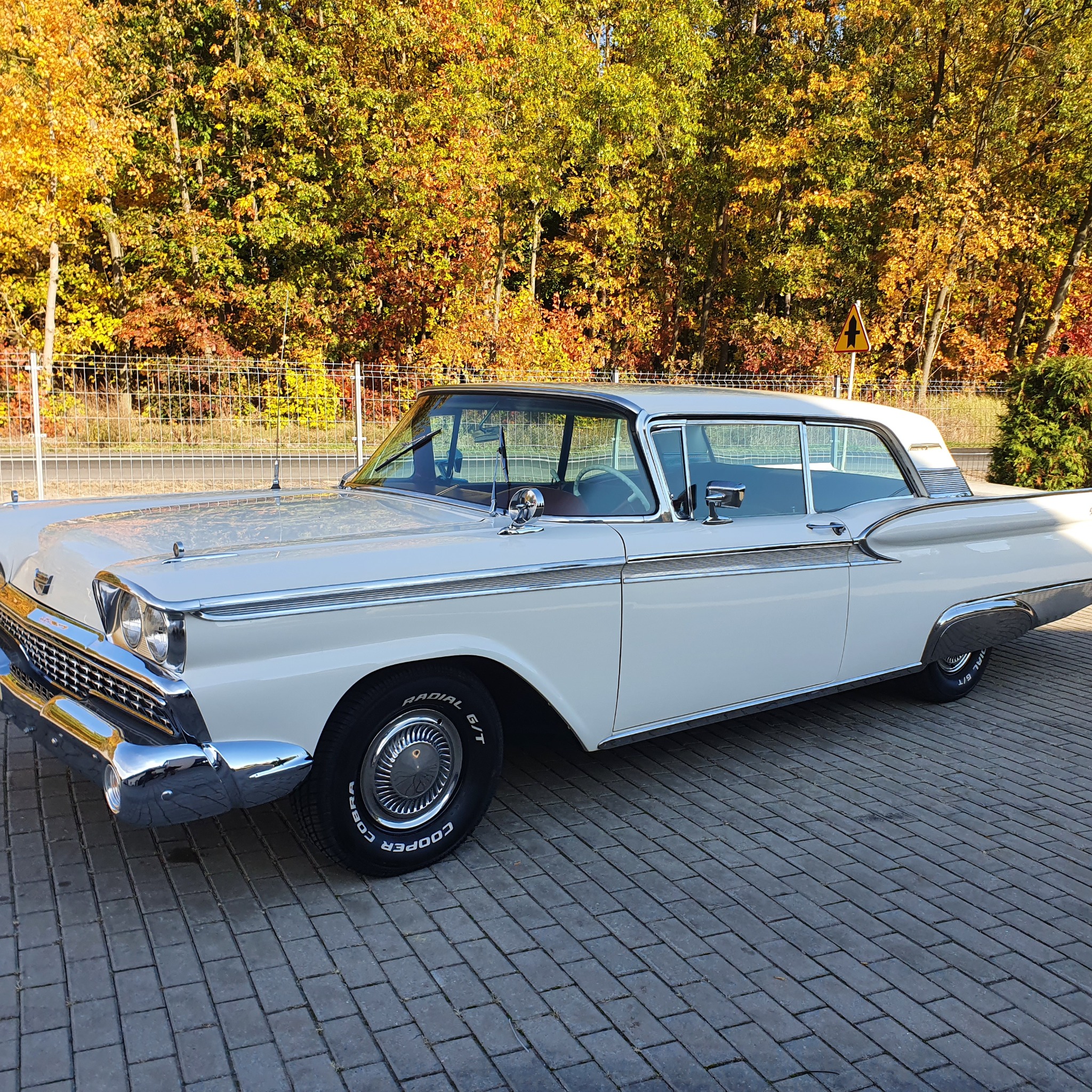 Biały, klasyczny samochód Ford Fairlane z 1959 roku zaparkowany na brukowanej nawierzchni, jesienne drzewa w tle.