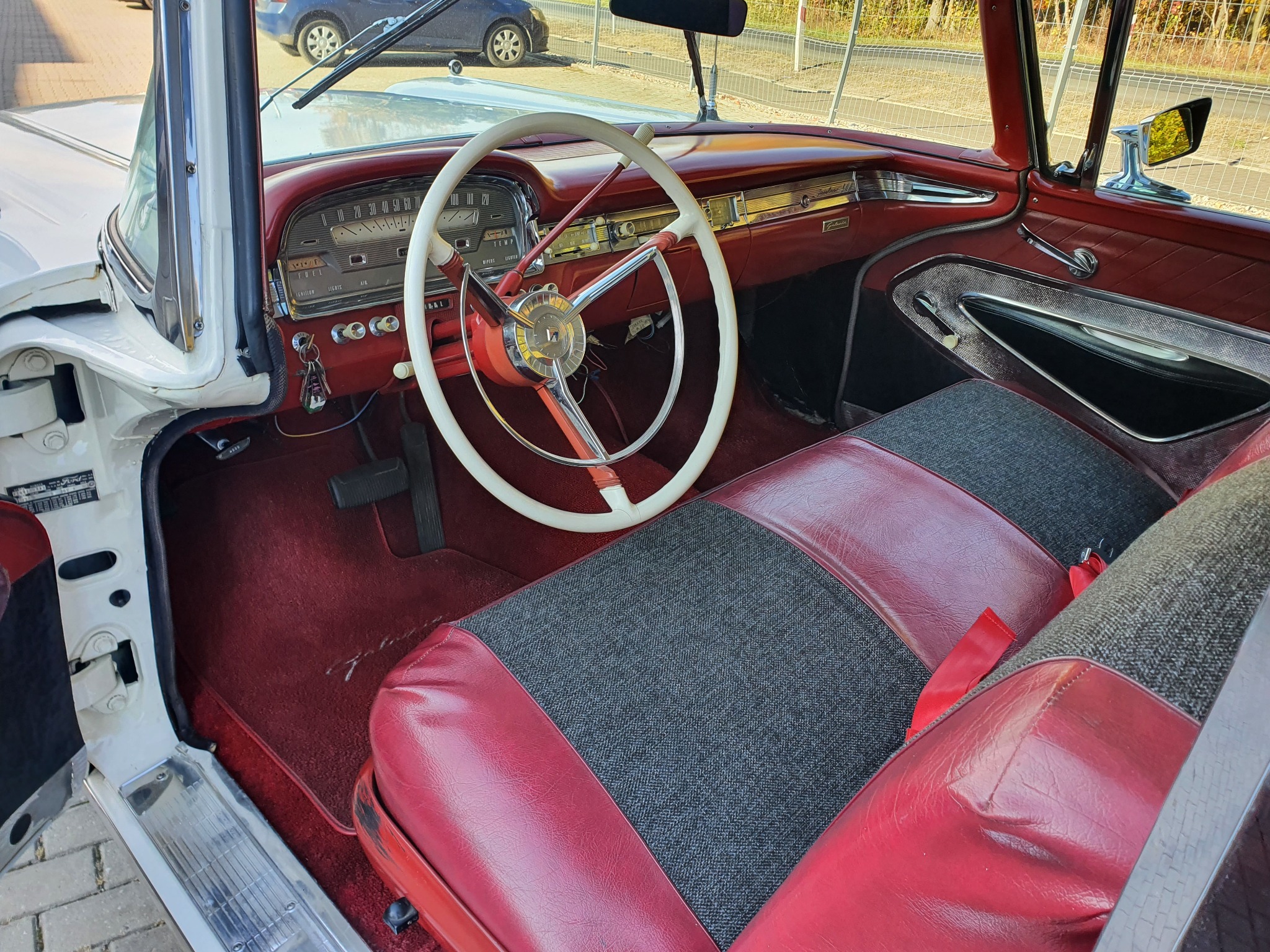 Wnętrze klasycznego samochodu z białą kierownicą, czerwonymi elementami deski rozdzielczej i siedzeniami w czerwono-szarej tapicerce. Widoczny fragment otwartych białych drzwi.