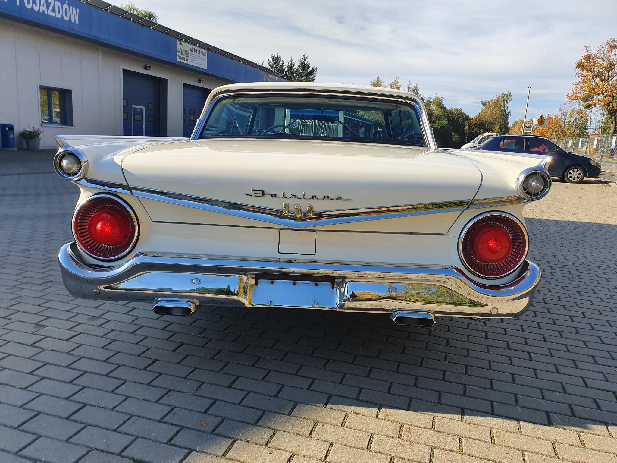 Tył klasycznego, kremowego Forda Fairlane 500 z chromowanymi detalami, widoczny na tle budynku warsztatu samochodowego z niebieskim dachem i brukowanym podjazdem.
