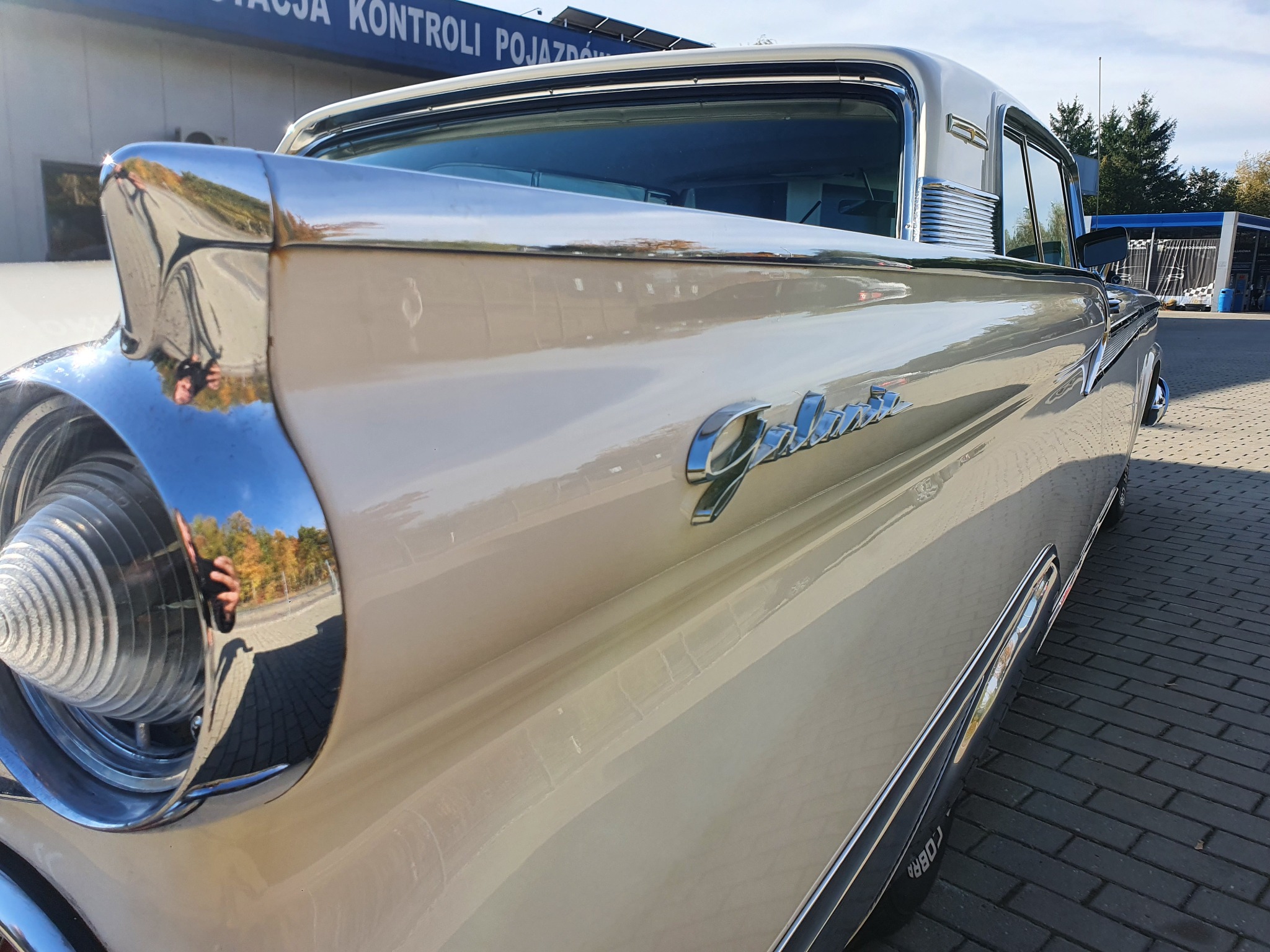 Fragment odrestaurowanego, kremowego Forda Galaxie z 1959 roku w Dąbrowie Chełmińskiej, z widocznym chromowanym reflektorem i napisem 'Galaxie', stojącego na kostce brukowej.