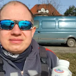 Autoportret pracownika w Jurzynie w okularach przeciwsłonecznych z odbiciem krajobrazu, na tle błękitnego samochodu dostawczego i zabudowań.