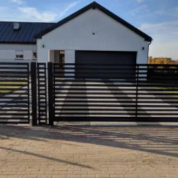 Nowoczesna, pozioma brama wjazdowa z metalowymi przęsłami, harmonizująca z architekturą domu w tle, z widocznym podjazdem wyłożonym kostką brukową.