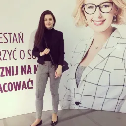 Kobieta w czarnym żakiecie i szarych spodniach pozuje na tle plakatu z wizerunkiem kobiety w okularach i marynarce w kratę, z widocznymi fragmentami tekstu reklamowego.