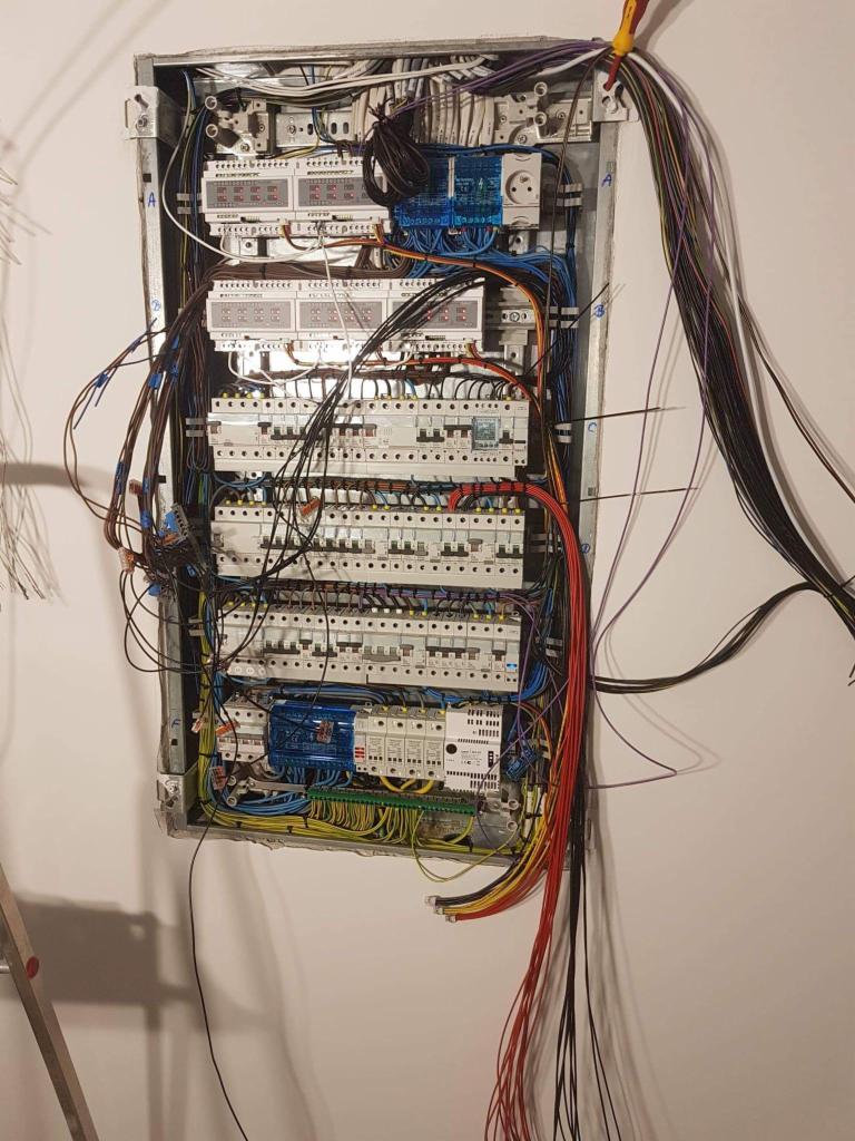 Otwarta skrzynka rozdzielcza z widocznymi bezpiecznikami, przewodami o różnych kolorach i elementami sterującymi, zamontowana na białej ścianie, z częściowo widoczną drabiną po lewej stronie.