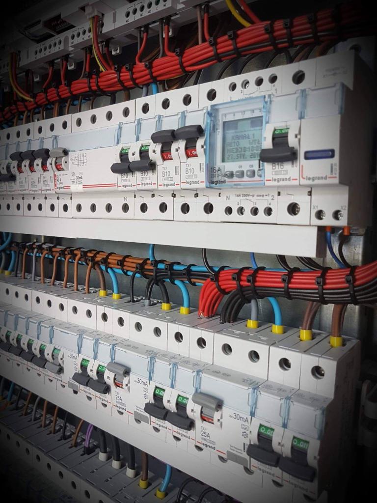 Rozdzielnica elektryczna z zainstalowanymi bezpiecznikami, przewodami w różnych kolorach, widoczne oznaczenia i etykiety, jeden z bezpieczników z wyświetlaczem LCD.