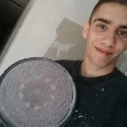 Uśmiechnięty mężczyzna trzyma w dłoni szlifierkę do gładzi na tle ściany ze śladami niedawnego szpachlowania, widoczne drobne zabrudzenia na ubraniu.