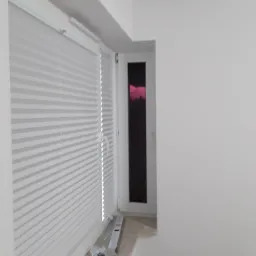 Białe, plisowane rolety okienne zamontowane w oknie, widoczna poziomica i akcesoria montażowe na parapecie, ściany pomalowane na biało.