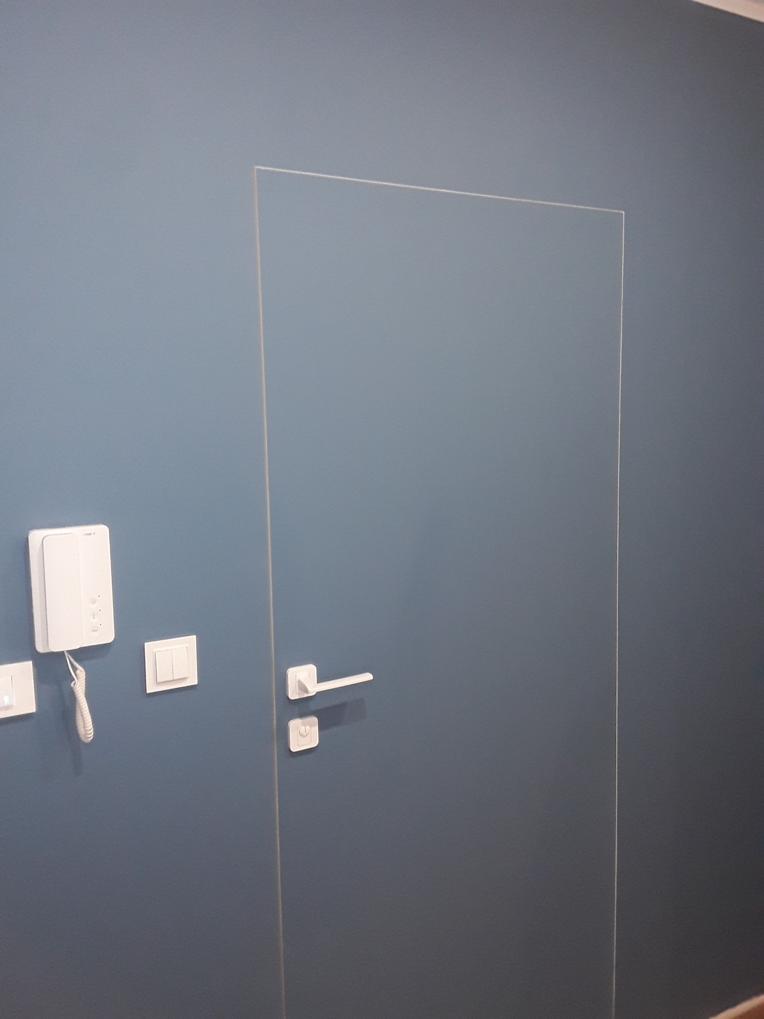 Minimalistyczne wnętrze z niebieską ścianą, białą, bezramową, szklaną furtką z białą klamką, białym domofonem i włącznikiem światła.