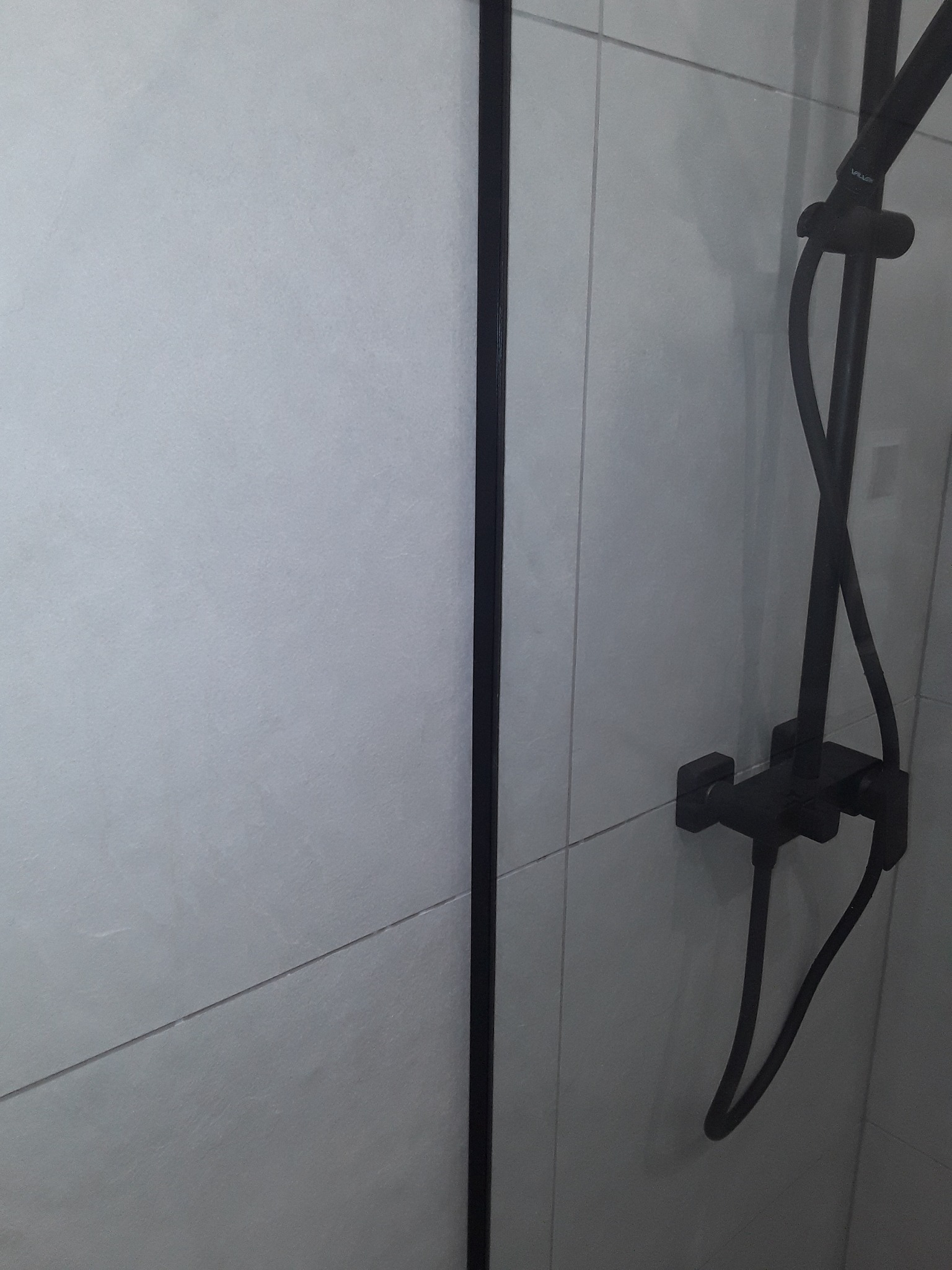 Szara łazienka z czarną kabiną prysznicową i armaturą, widok na kafelki i baterię prysznicową.