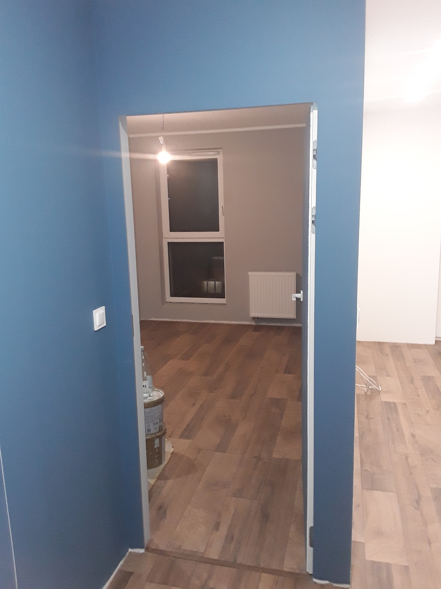 Widok przez otwarte drzwi na świeżo pomalowane wnętrze: błękitne ściany w przedpokoju i szare ściany w pokoju, drewniana podłoga, biały grzejnik i puszki z farbą przy progu.