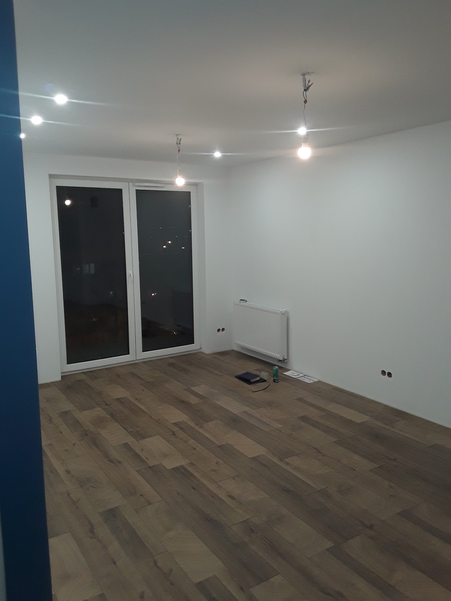 Pusty pokój po remoncie z panelami podłogowymi w odcieniach brązu, białymi ścianami, grzejnikiem i widocznymi przewodami oświetleniowymi zwisającymi z sufitu.