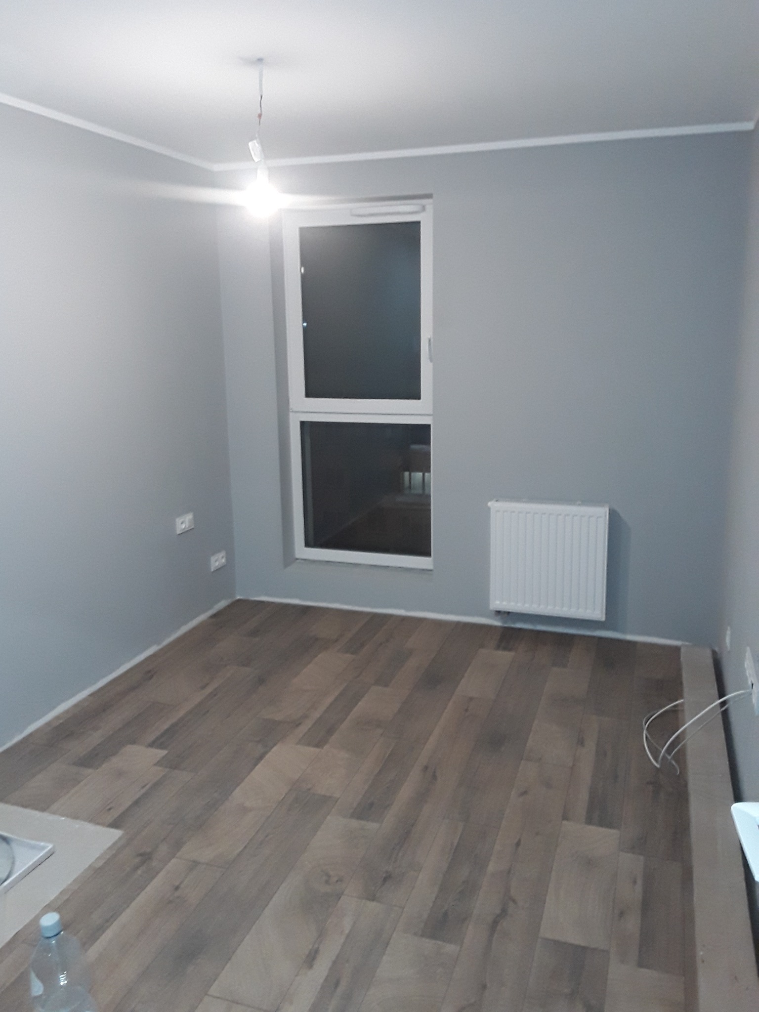 Pusty pokój po remoncie: szare ściany, panelowa podłoga, białe okno i grzejnik, widoczne gniazdka elektryczne i wisząca żarówka.