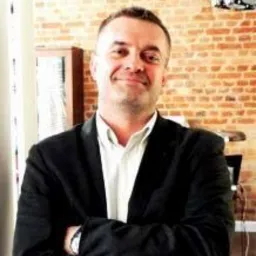 Portret uśmiechniętego mężczyzny w garniturze, stojącego na tle ceglanej ściany. Widoczne oświetlenie sufitowe i fragment biurka.