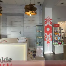 Wnętrze salonu kosmetycznego z ladą recepcyjną, dekoracyjnymi lampami z tektury, witrynami z kosmetykami i ścianą ozdobioną folkowym motywem kwiatowym.