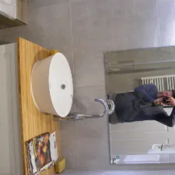 Nowoczesna umywalka nablatowa zamontowana na blacie z jasnego drewna, z widocznym odbiciem fotografa w lustrze, szare płytki na ścianie i biała szafka pod umywalką.