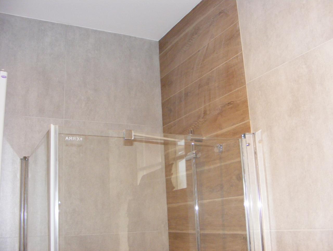 Szklana kabina prysznicowa z chromowanymi elementami, przylegająca do ściany wyłożonej szarymi płytkami oraz fragmentu ściany z płytkami imitującymi drewno.