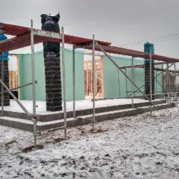 Wznoszenie surowego stanu domu z prefabrykatów na fundamentach w zimowej scenerii; widoczne rusztowania, zabezpieczone filary i zielone płyty konstrukcyjne.