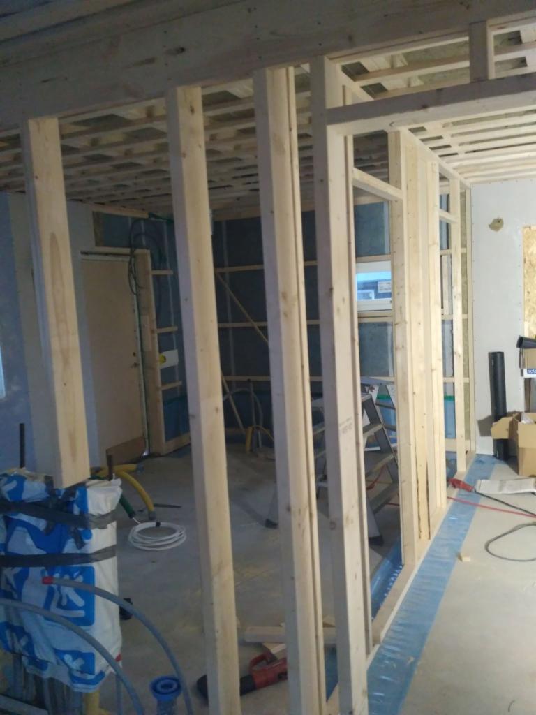 Wnętrze w trakcie budowy: widoczne drewniane ramy ścian działowych, sufit z belek, instalacje w podłodze i drabina w tle.