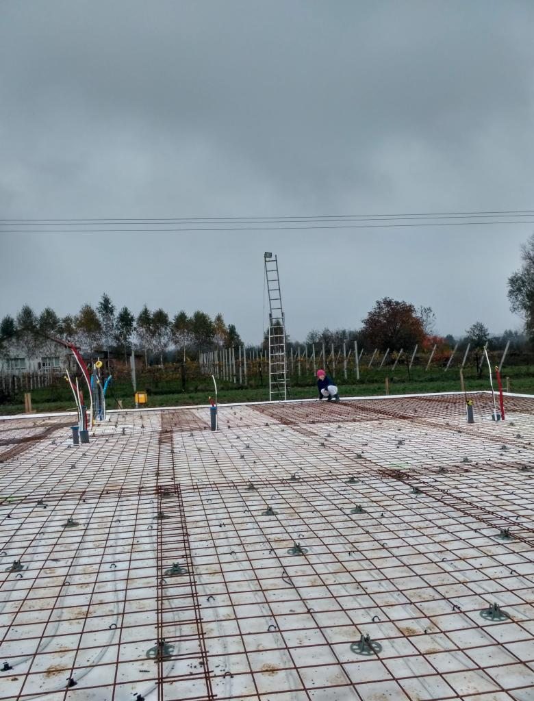 Przygotowanie fundamentów pod budynek: siatka zbrojeniowa na izolacji, wystające rury instalacyjne, drabina w tle, pochmurny dzień.