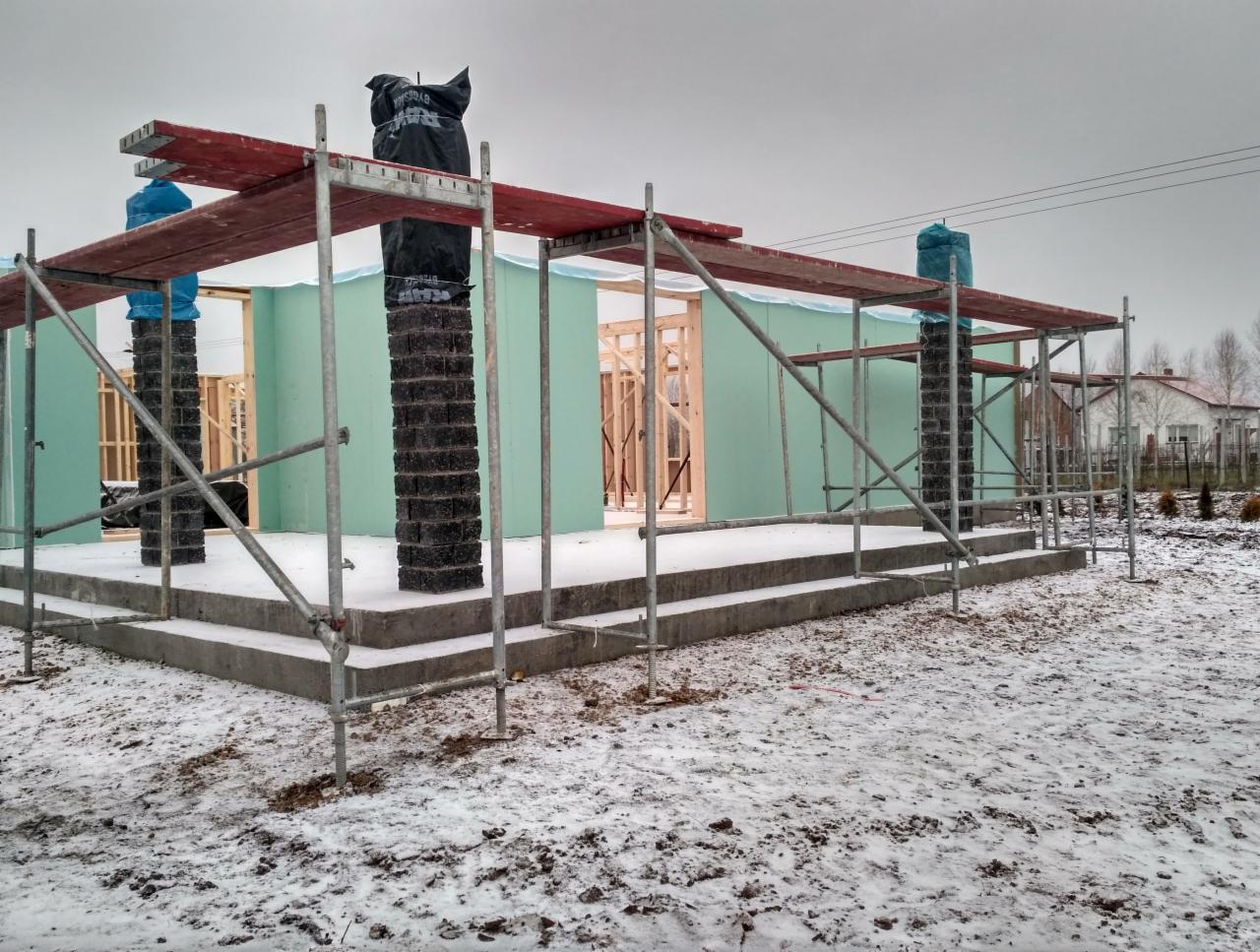 Wznoszenie surowego stanu domu z prefabrykatów na fundamentach w zimowej scenerii; widoczne rusztowania, zabezpieczone filary i zielone płyty konstrukcyjne.