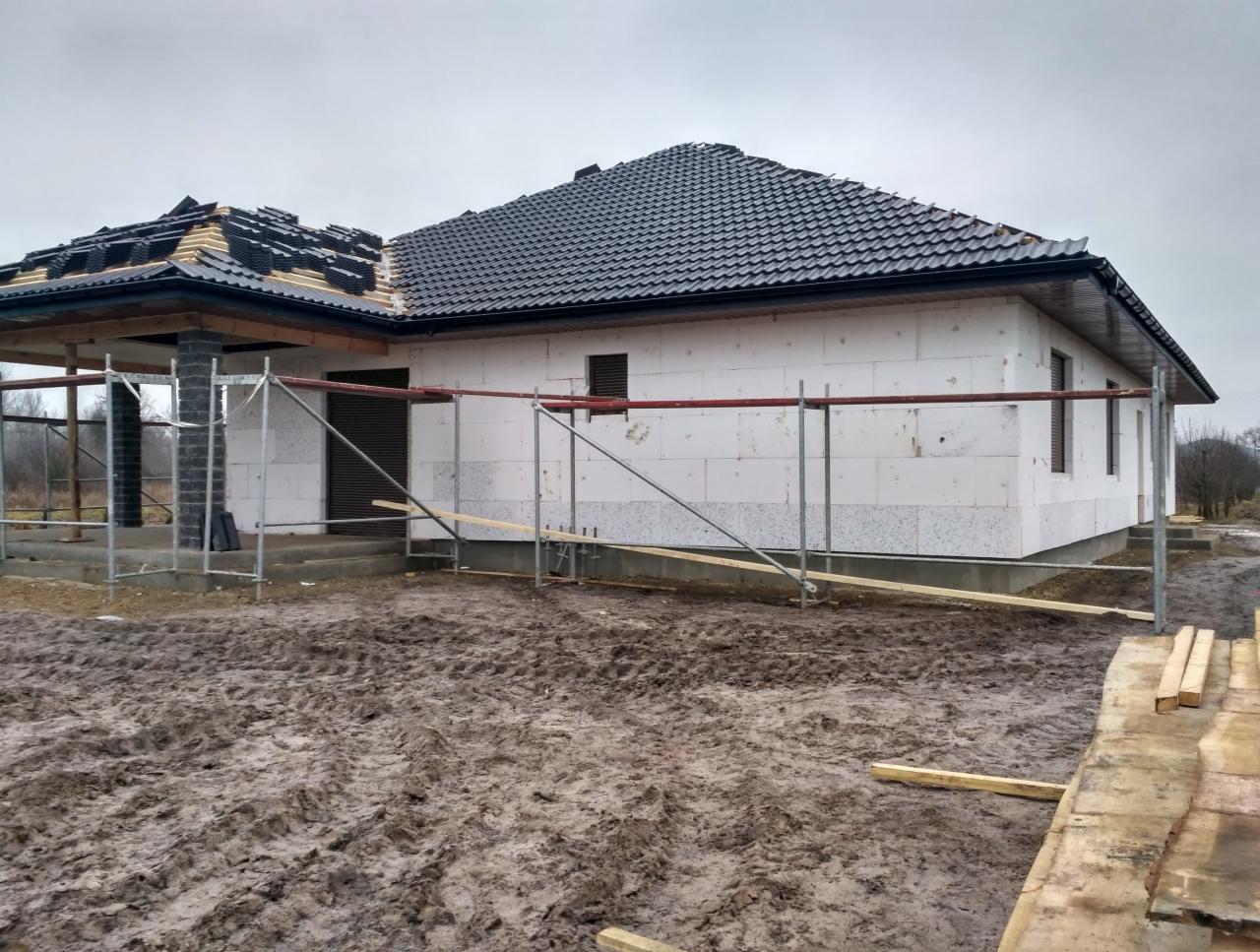 Budowa domu: stan surowy otwarty z częściowo ułożonym czarnym dachem z dachówki, ściany z białych bloczków, rusztowania wokół budynku, błotniste podłoże.