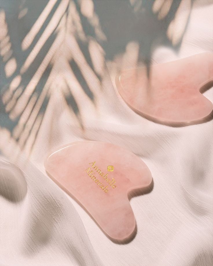 Dwa różowe kamienie Gua Sha 'Annabelle Minerals' na jedwabnej tkaninie w miękkim świetle z cieniem liści. Zdjęcie produktowe kosmetyków.