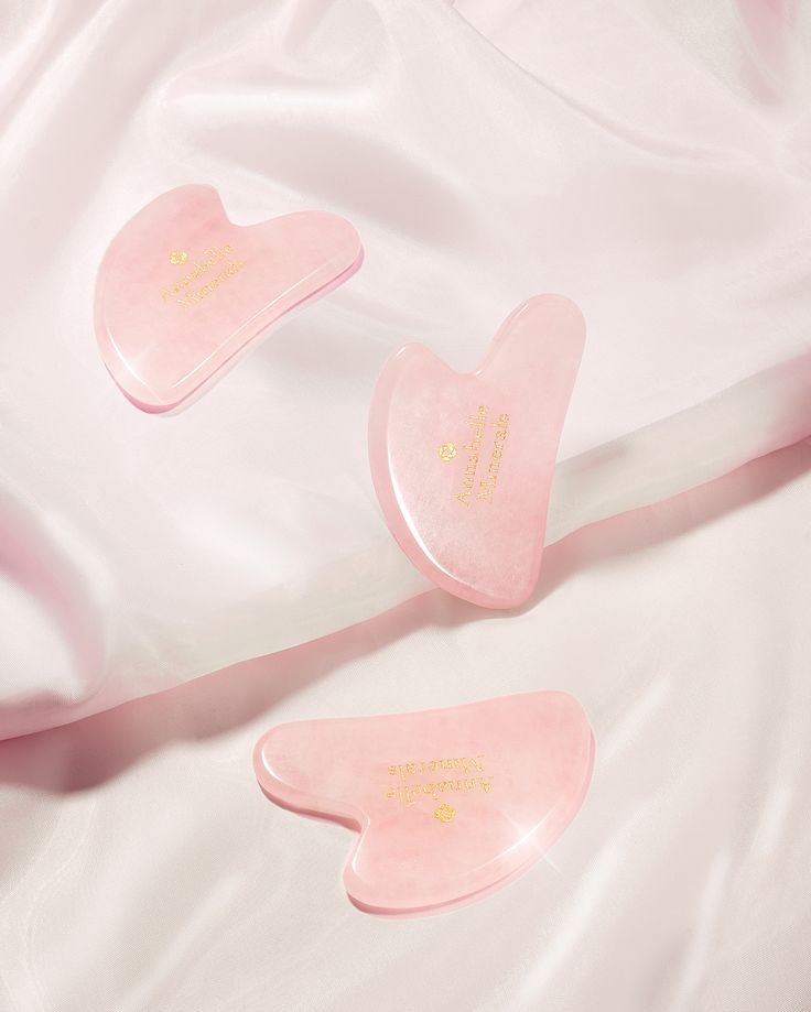 Trzy różowe płytki Gua Sha z napisem na jedwabnej, jasnoróżowej tkaninie. Produkt do pielęgnacji skóry w minimalistycznej aranżacji, z delikatnym oświetleniem.