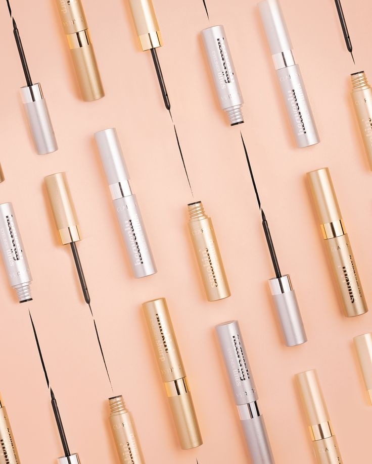 Stylizowany packshot: złote i srebrne eyelinery Clarins, ułożone pod kątem na brzoskwiniowym tle, z aplikatorami i śladami tuszu, prezentacja kosmetyków do makijażu.