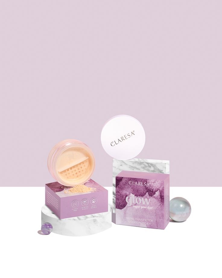 Zdjęcie pudru sypkiego Claresa Glow w otwartym opakowaniu, z pudełkiem i kulami na białym podeście, na fioletowym tle. Packshot kosmetyczny.